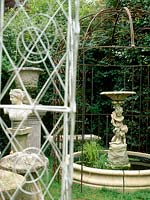 Cherub fountain with two playful cherubs, stone bench, garden statue and ornate vase on plinth viewed through antique pergola.