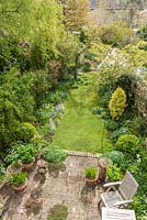 Aerial view of long, narrow, town garden in spring with informal lawn and mixed borders. Reclaimed brick patio with hostas and agapanthus in containers.