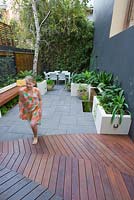 Young girl walks to timber decking in inner city courtyard with small dining area and back fence covered with Phylostachys nigra, black bamboo. Concrete planters with various shade loving plants seen. 