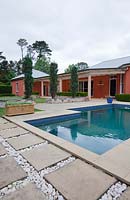 Swimming pool at back of house featuring three conifers a trough pot and concrete stepping stones with white quartz pebble insets.