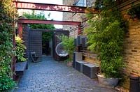 Alleyway beside house with wall lighting, bamboo in container galvanised bath, steel girder pergola and wooden fence with open gate at the end. Brick flooring. Metal storage boxes hung on a rack on the wall.

