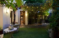 Courtyard garden at night. Cafe table and chairs between grape vine in a large container. Two childrens' swings hung from unseen steel girder pergola covered in climber, open front door, planting of phyllostachys bamboo.  