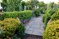 Paving path between borders planted with a tapestry of shrubs including golden spirea, bay, griselinia and edging of dianthus. Sculptural ornaments including stained glass panels. 