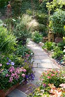 Paving path edged with brick between borders planted with phlomis, stipa, euonmyus and annuals in containers. Rusted steel obelisk, found wooden blocks. Ornamentation.  