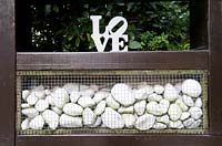 Wooden frame with white pebbles inset and retained by wire mesh with Love ornament on top.