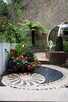 Low brown painted wall retaining raised beds, plants in containers including salvia and agapanthus, tree fern Dicksonia antarctica, high neighbouring boundary wall and seating inside a large round pod. Inlaid pebble mosaic hard landscape flooring