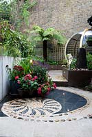 Low brown painted wall retaining raised beds, plants in containers including salvia and agapanthus, tree fern Dicksonia antarctica, high neighbouring boundary wall and seating inside a large round pod. Inlaid pebble mosaic hard landscape flooring