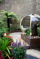 View of garden with brown painted low retaining walls and raised beds with colourful planting including salvia and agapanthus with a tree fern, high neighbouring wall, change of levels, hard landscaping, lighting posts and a large round wooden and perspex pod with seating and a table. Outdoor room. 