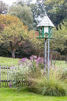 Wooden bird feeder standing over scene with perennials and grasses next to wooden picket fence. Pasture and apple trees in the background