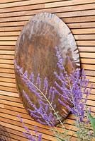 Copper hammered disc on wall in garden with perovskia flowers. Ben De Lisi House and Garden, London