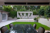 View from a cabana to a garden wall featuring an inset with a zig zag wire pattern trellis and a white flowering Stephanotis floribunda growing on it, stone paving steps and a Buffalo grass lawn.