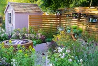 Side view across garden to pink-painted wooden shed. Large circular earthenware water feature with waterlilies on gravel, with raised beds surrounding it, and garden tools and vases of cut flowers on a wooden table at the back of the garden. Plants in the foreground include Rosa 'Blush Noisette',  Euphorbia, Astrantia major 'Pink Pride', and  chocolate cosmos - Cosmos 'Chocamocha'. RHS Hampton Court Palace Flower Show 2016