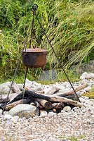 The Viking Cruises Scandinavian Garden, detail of rusted cooking pot on metal tripod over charred logs, on pebble and shingle , with wildflower meadow behind. RHS Hampton Court Flower Show 2016