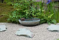 Detail of a 'Japanese Summer Garden' designed by Saori Imoto showing the gravel path set with granite stepping stones, on which sits a stone bowl for water with a bamboo spout and wooden ladle. Planting includes moss, nandina, ferns and iris. A low bamboo fence borders the garden on one side. Hampton Court Flower Show, July 2016.