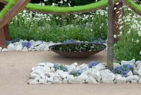 'The World Vision Garden', designed by John Warland, showing waves of mild steel on triangular supports, planted with lush green turf, punctuated by the vertical trunk of an ornamental pear tree and softened by the planting of ox-eye daisies. RHS Hampton Court Flower Show, July 2016.