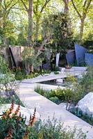 The Telegraph Garden, view of irregular geometric bronze fin sculptures, seating area with stone benches, limestone paths surrounded by stones, Isoplexus canariensis - Canary Island Foxgloves, Maytenus boaria, Schinus molle. RHS Chelsea Flower Show 2016. Designer: Andy Sturgeon - Sponsor: The Telegraph