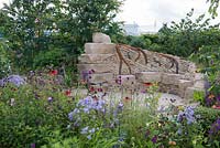 Dry stone seating area with steel tree root sculpture, herbaceous border in foreground with Cirsium rivulare, Echinacea and Campanula - Zoflora: Outstanding Natural Beauty, RHS Hampton Court Palace Flower Show 2016