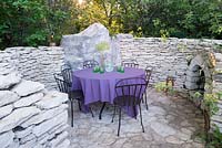 L'Occitan Garden, view a patio with black metal chairs, table with purple table cloth, glasses, jug and lanterns surrounded by curved sand stone fence with fire place.  RHS Chelsea Flower Show, 2016.
