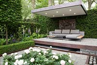 The Husqvarna Garden, view of outdoor living area on floating stone terrace with sofa and cushions, covered canopy with lighting, copper water rill, pleached Hornbeam, Taxus baccata and Buxus sempervirens hedge, Paeonia 'Elsa Sass', Angelica gigas and Digitalis purpurea. RHS Chelsea Flower Show 2016, Designer: Charlie Albone, Sponsors: Husqvarna