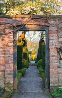 A gateway through a red brick wall into The Well Garden at Wollerton Old Hall Garden, Shropshire - photographed in April. Beyond the gate are clipped pyramidal yews and above it, a blossoming cherry tree
