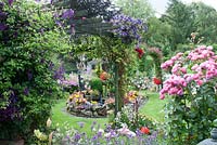 View of colourful small back garden with borders filled with tender bedding plants, perennials, Rosa 'Pomponella' and Clematis 'Jackmanii' on trellis and pond