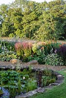 Sue O'Neill's garden at Field Cottage, Cirencester, Glos. Large pond with gravel edge and summery border behind
