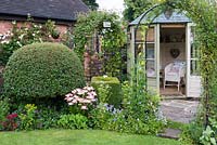 A cottage garden with summer house, clematis covered arch, topiary box and privet and mixed border with violas, lilies and geraniums.