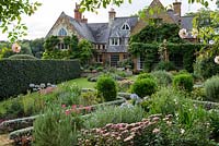 Coton Manor and a parterre with pink and blue planting, including Sedum 'Matrona', Caryopteris 'Heavenly Blue', Agapanthus 'Blue Moon', Cleome hassleriana 'Rose Deep', Knautia macedonica, Cuphea viscosissima and Gladiolus murielae.