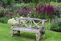 A wooden bench in front of a purple and blue autumn perennial border. Planting includes Clematis heracleifolia 'Cassandra', Salvia involucrata, Dahlia sherfii, Rosa 'Louise Odier', Sedum spectabile and 'Frosty Morn', Lobelia 'Tania' and 'Hadspen Purple'.