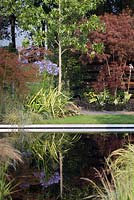 A dark refelctive pond and a fire pit in a contemporary garden with lawn, grasses, trees and perennials - July, RHS Tatton Park Flower Show, Cheshire