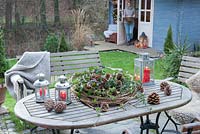 Garden table decorated with wreath of Clematis vines with Gaultheria procumbens, Hedera and cones, lanterns and pine cones

