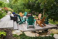 Brown wicker furniture, green and tan wooden Adirondack chairs on grey wooden patio deck with firepit in sloped backyard garden in summer