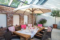 Table prepared for coffee time in a winter garden with brick stone wall and white parasol, Ficus carica and Pelargonium
