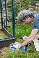 Man using cordless combi drill, with screwdriver attachment, to fix 'plastic wood' edging plank in place with brass screws - having first made a pilot hole using a hand drill: April, Spring.