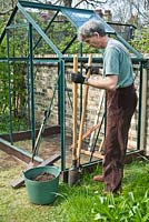 Man using post hole tongs to dig a hole, in which to insert a 'plastic wood' post to help secure the base of the greenhouse in place: April, Spring.