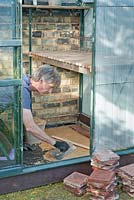 Man using a plastering trowell to smooth over a layer of sharp sand, over the top of a sheet of black geotextile membrane, on which to lay a floor of quarry tiles: April, Spring.