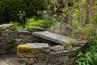 Curved stone wall seating area with succulents and naturalistic spring planting including Syringa - Lilac tree - Macmillan Legacy Garden - RHS Malvern Spring Show 2016. Designer: Mark Eveleigh, Sponsor: Macmillan Cancer Support