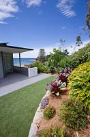 The back garden of a beach-side Australian house built on a steep site. Artificial lawn and a raised bed featuring Alcantareas and Philodendron 'Xanadu' is seen.