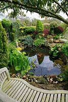 Fish pond with a backdrop of evergreens, marginal plants,  pond side seats and wires stretched over the surface to deter herons. Design: David Green and Elaine MacKenzie.