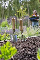Garden trowel and hand fork in a raised bed with young girl carrying tray of flowers in background