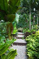 A stepped gravel path with timber risers leading to a garden bench flanked by a thickly planted lush garden.