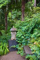 A Balinese carved stone lantern sits on a large black timber plinth next to a simple timber bench at the end of a pea gravel path surrounded by a thick planting of ferns and other shade loving plants.