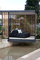 A black steel framed garden pavilion sits on top of a water feature, with cedar timber batten 