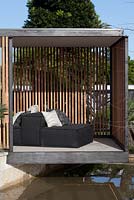 A black steel framed garden pavilion sits on top of a wall above a water feature, with lounge seats with cushions