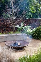 A fire pit with on a paved area in front of a timber garden bench surrounded by a planting of mixed plants and grasses