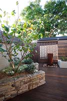 Inner city courtyard with featuring a raised bed with stone stack wall with a Cercis  tree,a freestanding ceramic water feature, a rusty corten decorative screen and a timber slat panel wall