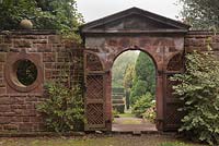 A view through a pseudo-classical style red sandstone lattice gateway to conifers in garden beyond - October, Burton Manor, Cheshire