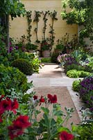 Stone and gravel pathway leading to cordoned pear trees in a courtyard garden growing flowers, fruit and vegetables. Plants include Lavandula Regal Splendour, Artichoke, Ruby chard and Taxus baccata