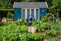 A summer-house in a vegetable garden planted with strawberries, potatoes, lettuce, broad beans and asparagus. A rusted bird sculpture in the centre