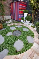 A corten steel edged firepit surrounded by round stone steps and planted with Violia hederacea, Australian native violets. Area is surrounded by sandstone edging and a colourful striped shed with white pots beside are also seen